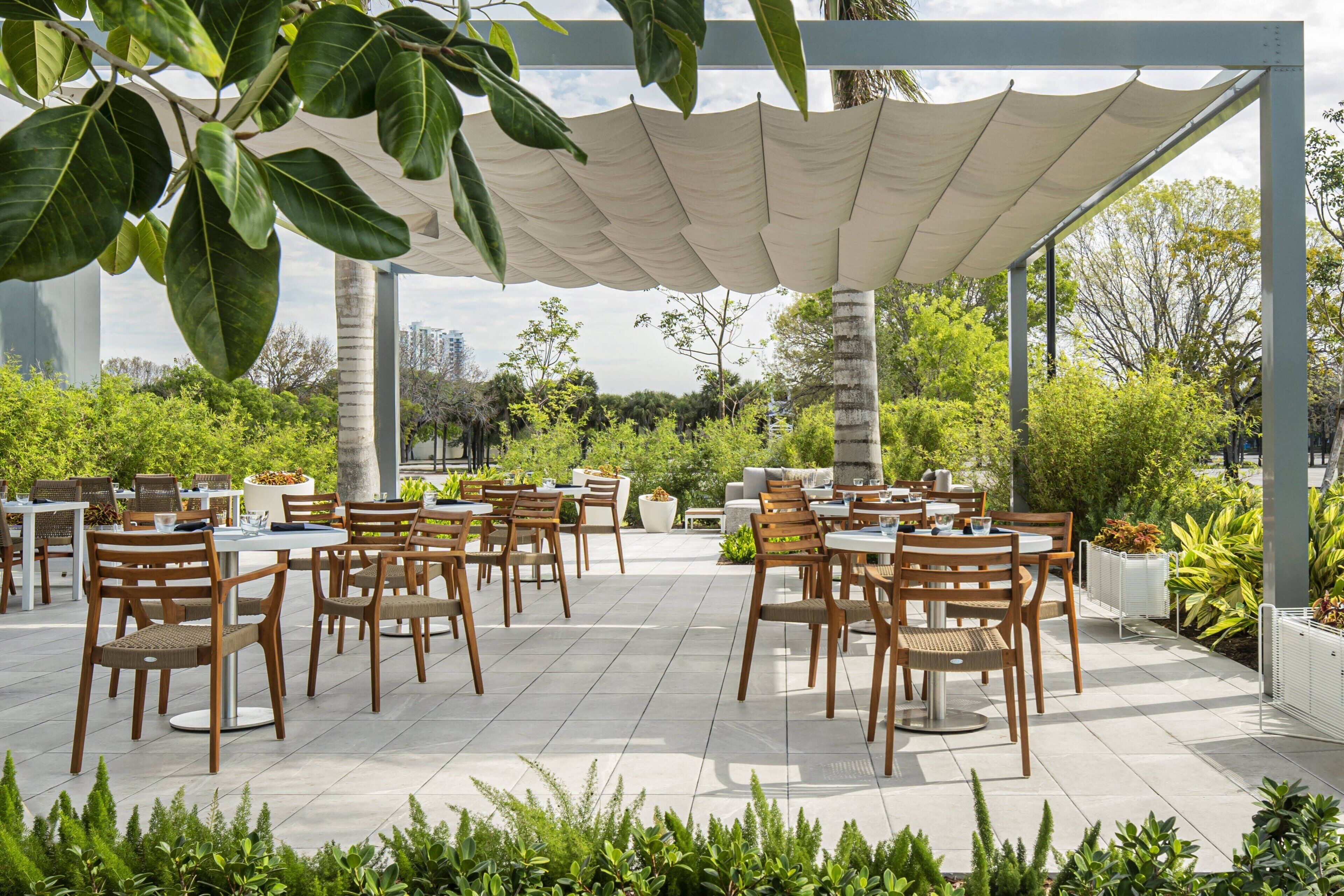 An outdoor patio with multiple tables and chairs, surrounded by greenery and a covered area with a white canopy.