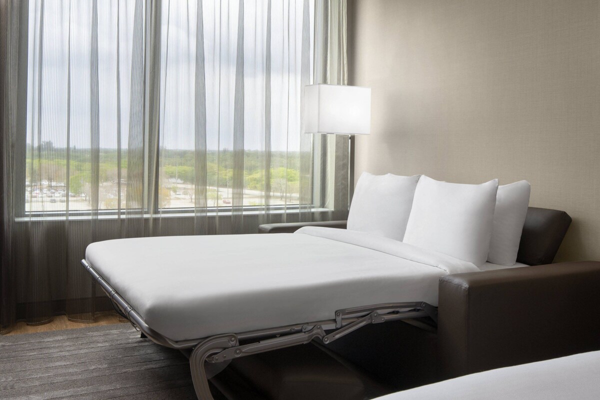 A hotel room with a bed, a bedside table, a lamp, and a view of the outdoors through sheer curtains.