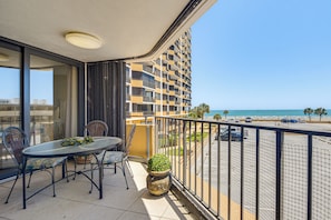 A balcony with a table and chairs, a potted plant, and a view of the beach and ocean.