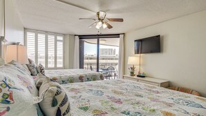 Une chambre d’hôtel avec deux lits, un ventilateur de plafond, une télévision et un balcon donnant sur des immeubles.