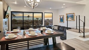 A modern dining area with a large table set for four, a chandelier, and a scenic view through large windows. - Newly remodeled home with breathtaking ocean view. (Newport Beach)