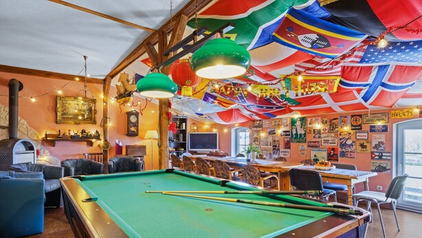 Mesa de bilhar com tacos, rodeada por poltronas de couro e um espaço de bar com relógio e diversas decorações.