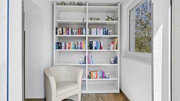 A room with a white bookshelf filled with books, a white chair, and a window with a view of trees.