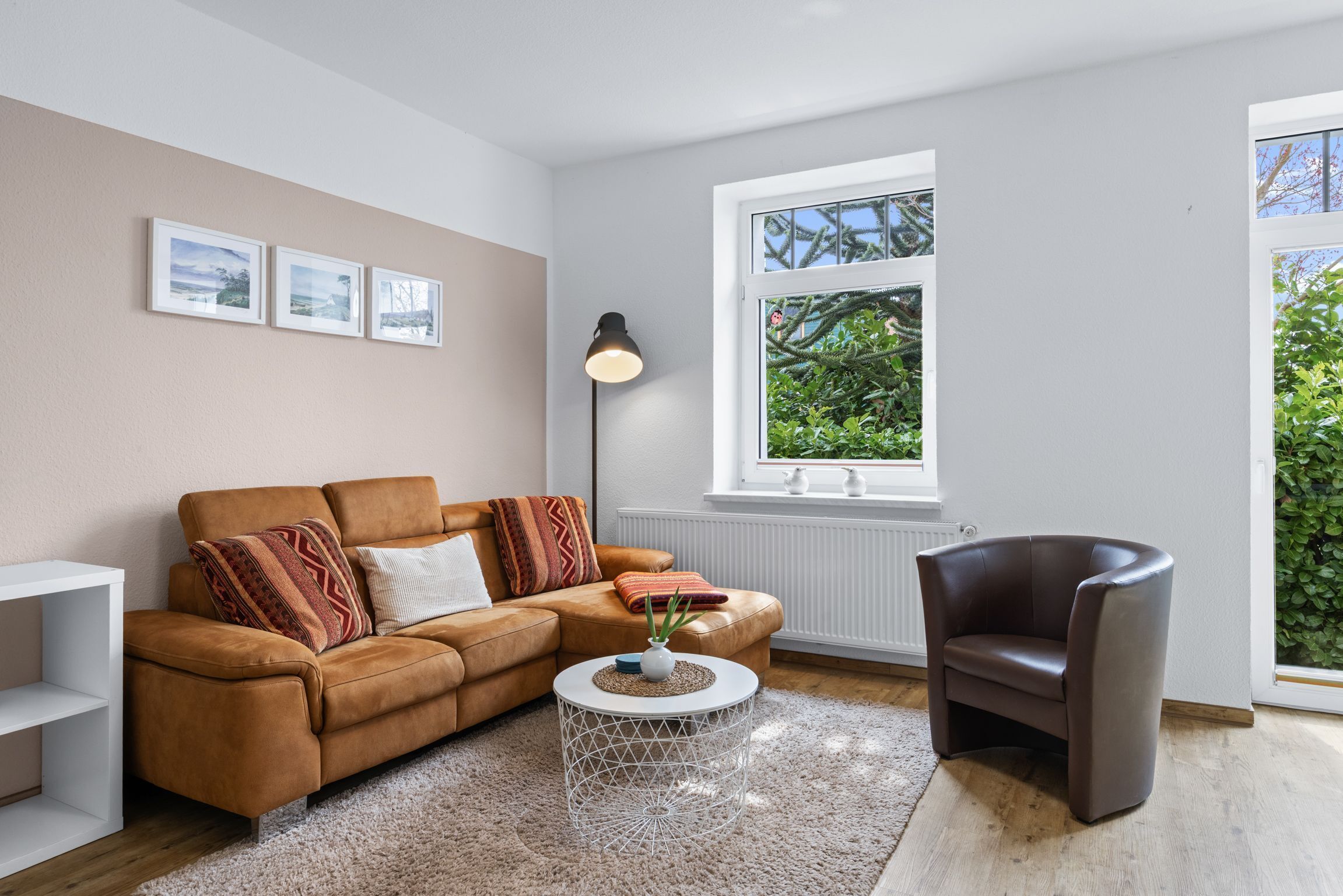 A modern living room with a brown leather sofa, a white side table, a black floor lamp, and a window with a view of greenery.