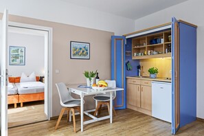 Ein kleines Zimmer mit Bett, Esstisch mit Stühlen, einer Küchenzeile und einer Tür zu einem weiteren Raum.