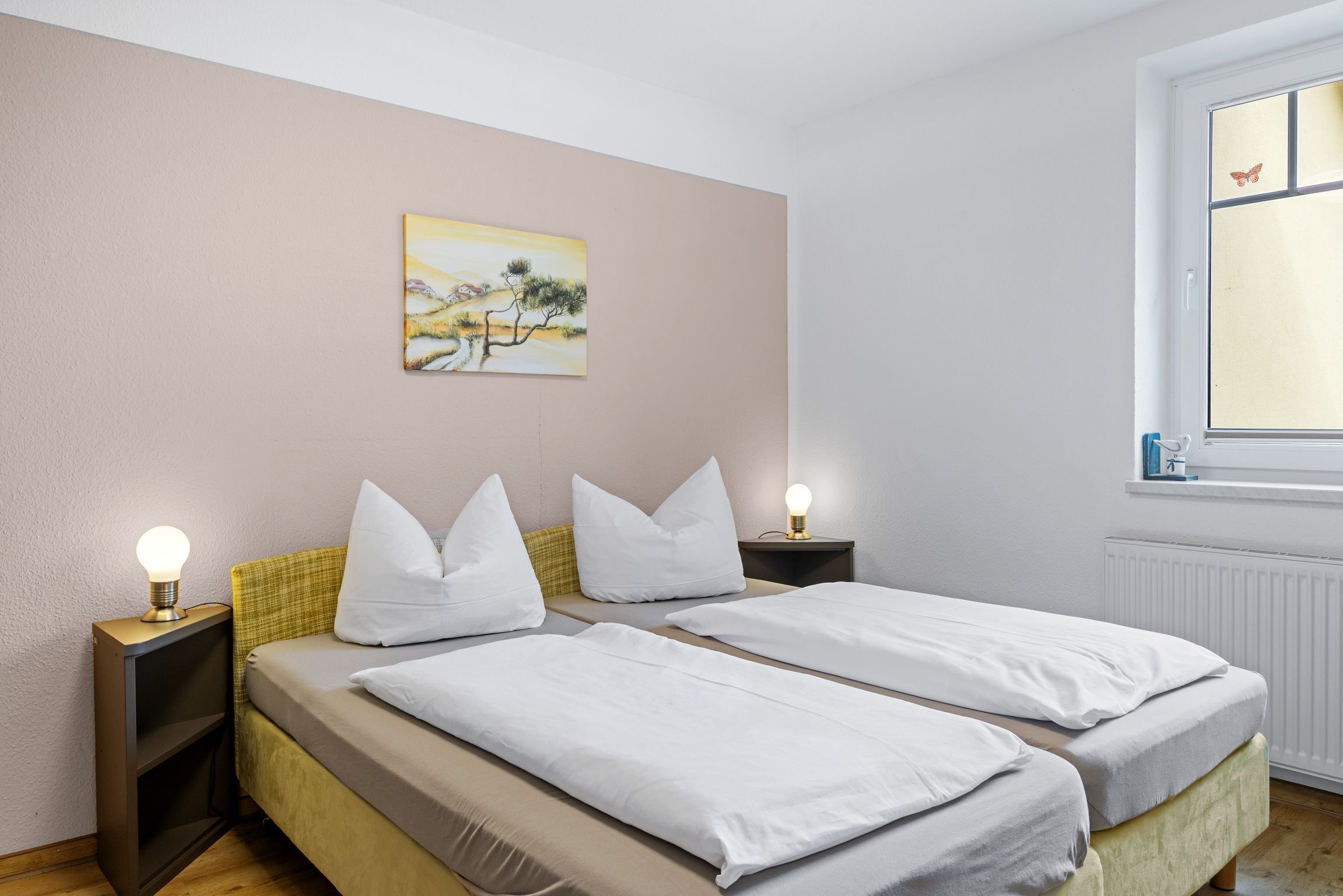 Een slaapkamer met twee bedden, een nachtkastje met een lamp, een schilderij aan de muur en een raam met uitzicht.