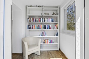 Ein Zimmer mit einer weißen Bücherwand, einem weißen Stuhl und einem Fenster mit Blick auf Bäume.