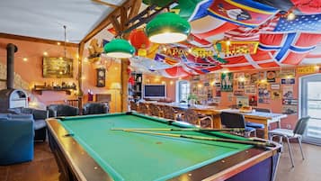 Une table de billard, avec des queues de billard, entourée de fauteuils en cuir et d’un coin bar agrémenté d’une horloge et de divers objets de décoration.