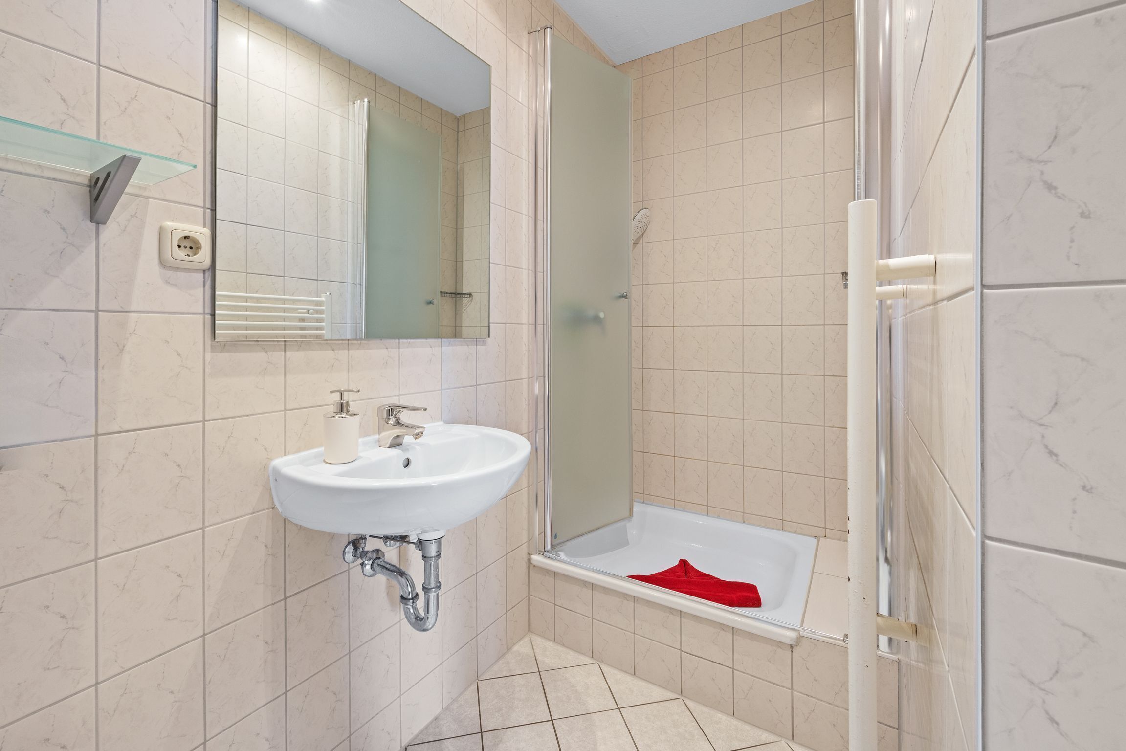 Un baño con lavamanos blanco, un espejo, cabina de ducha de vidrio y una toalla roja en el suelo.