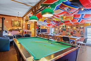 Une table de billard, avec des queues de billard, entourée de fauteuils en cuir et d’un coin bar agrémenté d’une horloge et de divers objets de décoration.