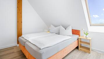 A modern bedroom with a sloped ceiling, a bed with grey bedding, a small wooden nightstand, and a window with a view of the sky.