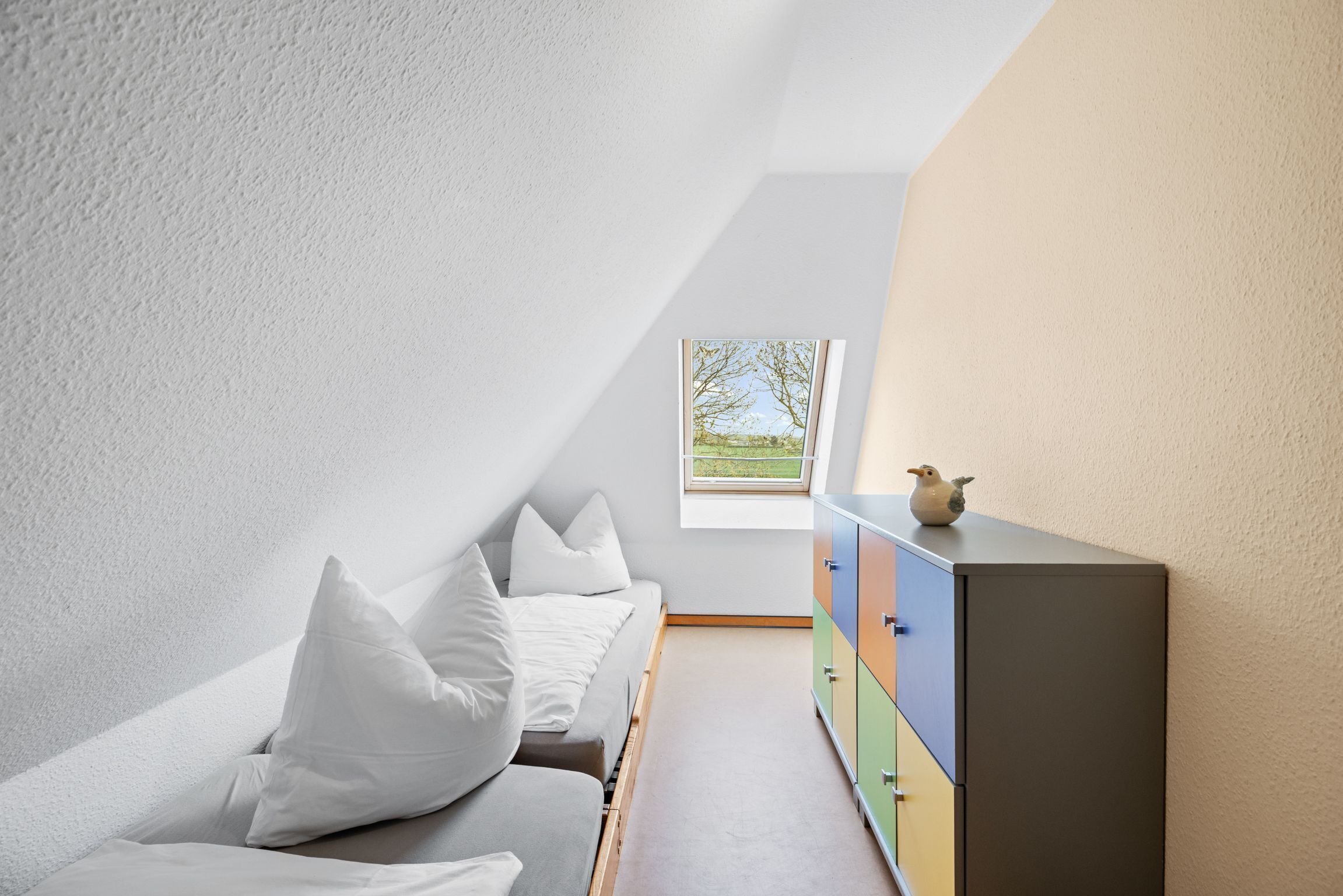 A room with a sloped ceiling, a bed with white bedding, a colorful dresser, and a window with a view of greenery.