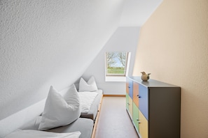 A room with a sloped ceiling, a bed with white bedding, a colorful dresser, and a window with a view of greenery.