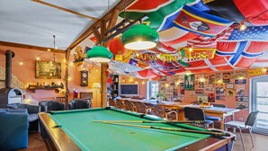 A pool table with pool cues, surrounded by leather chairs and a bar area with a clock and various decorations.
