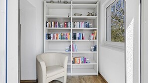 A room with a white bookshelf filled with books, a white chair, and a window with a view of trees.