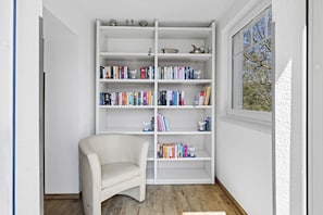 A room with a white bookshelf filled with books, a white chair, and a window with a view of trees.