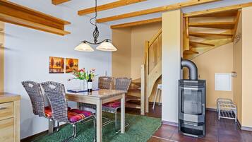 A dining area with a wooden table, wicker chairs, and a green rug. A wood-burning stove is visible in the corner.