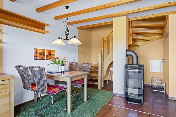 Un coin repas avec une table en bois, des chaises en osier et un tapis vert. Un poêle à bois est visible dans un coin.