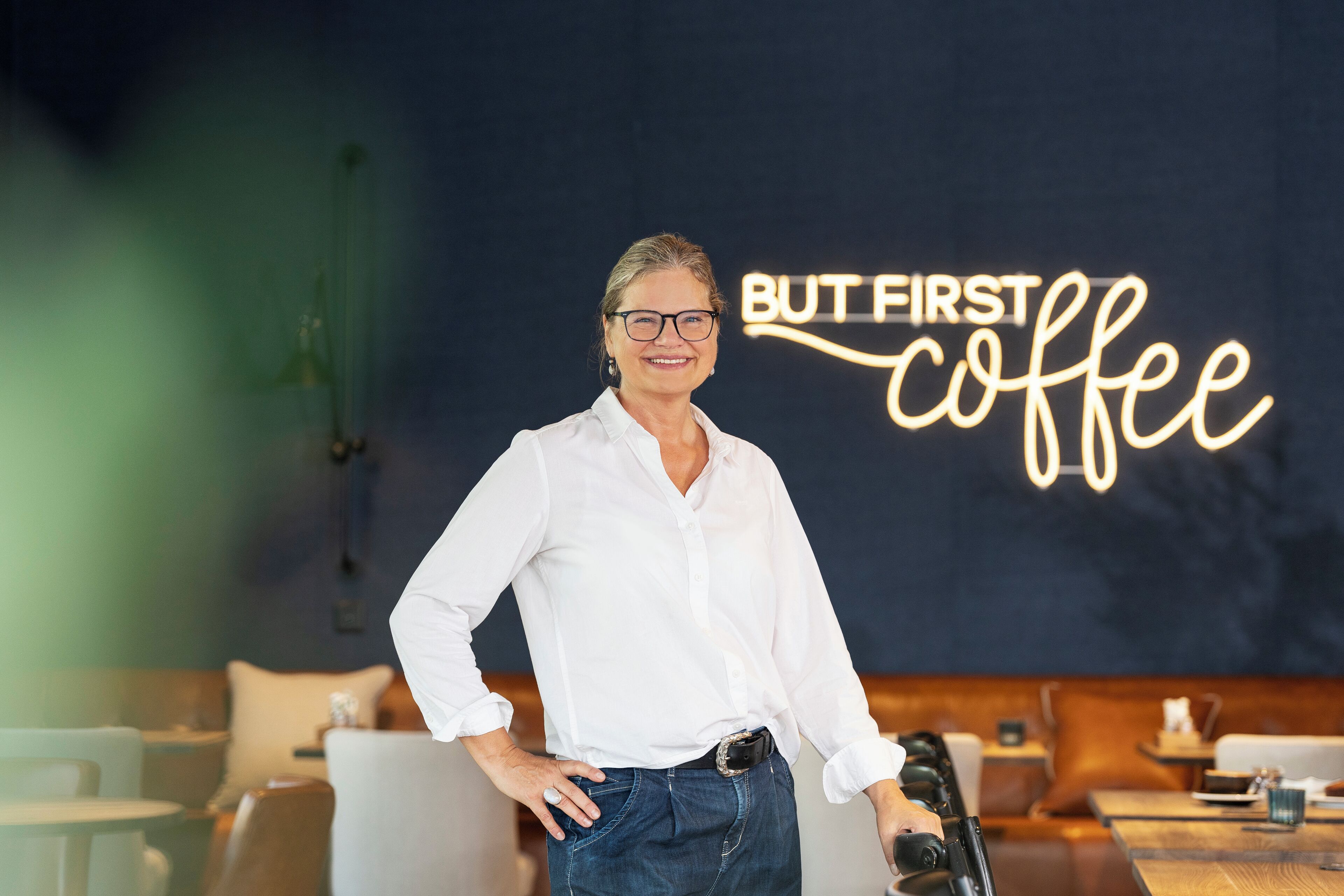 A person standing in a modern interior with a neon sign reading "BUT FIRST coffee" on the wall.