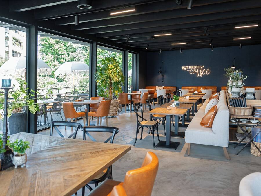 A modern cafe with wooden tables and chairs, a sofa, and large windows offering a view of the outdoors.