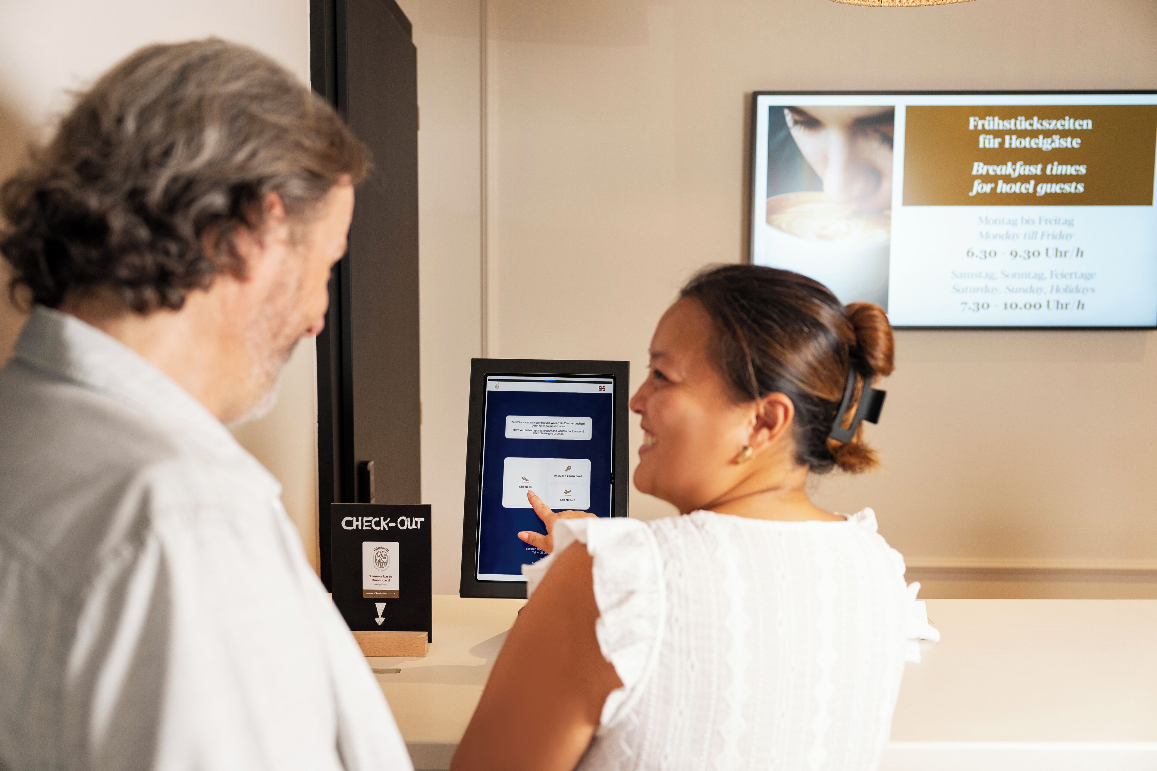 A hotel reception area with a digital check-in kiosk and a sign indicating breakfast times for hotel guests.