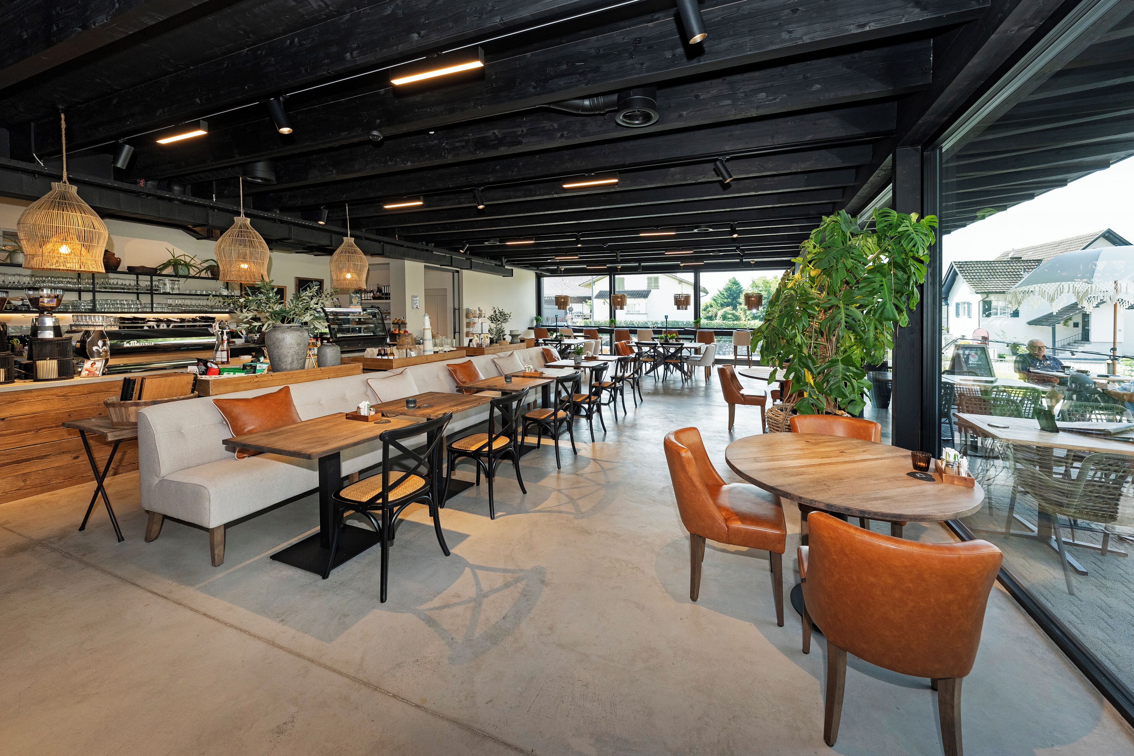 Una cafetería moderna con una barra larga, zona de mesas y grandes ventanales que ofrecen vistas al exterior.