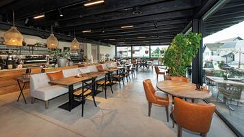 Una cafetería moderna con una barra larga, zona de mesas y grandes ventanales que ofrecen vistas al exterior.