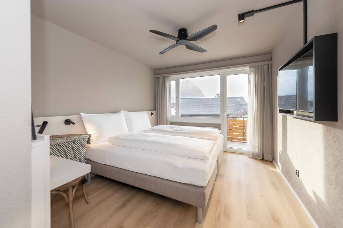 A modern bedroom with a bed, a desk, a ceiling fan, and a large window with curtains.