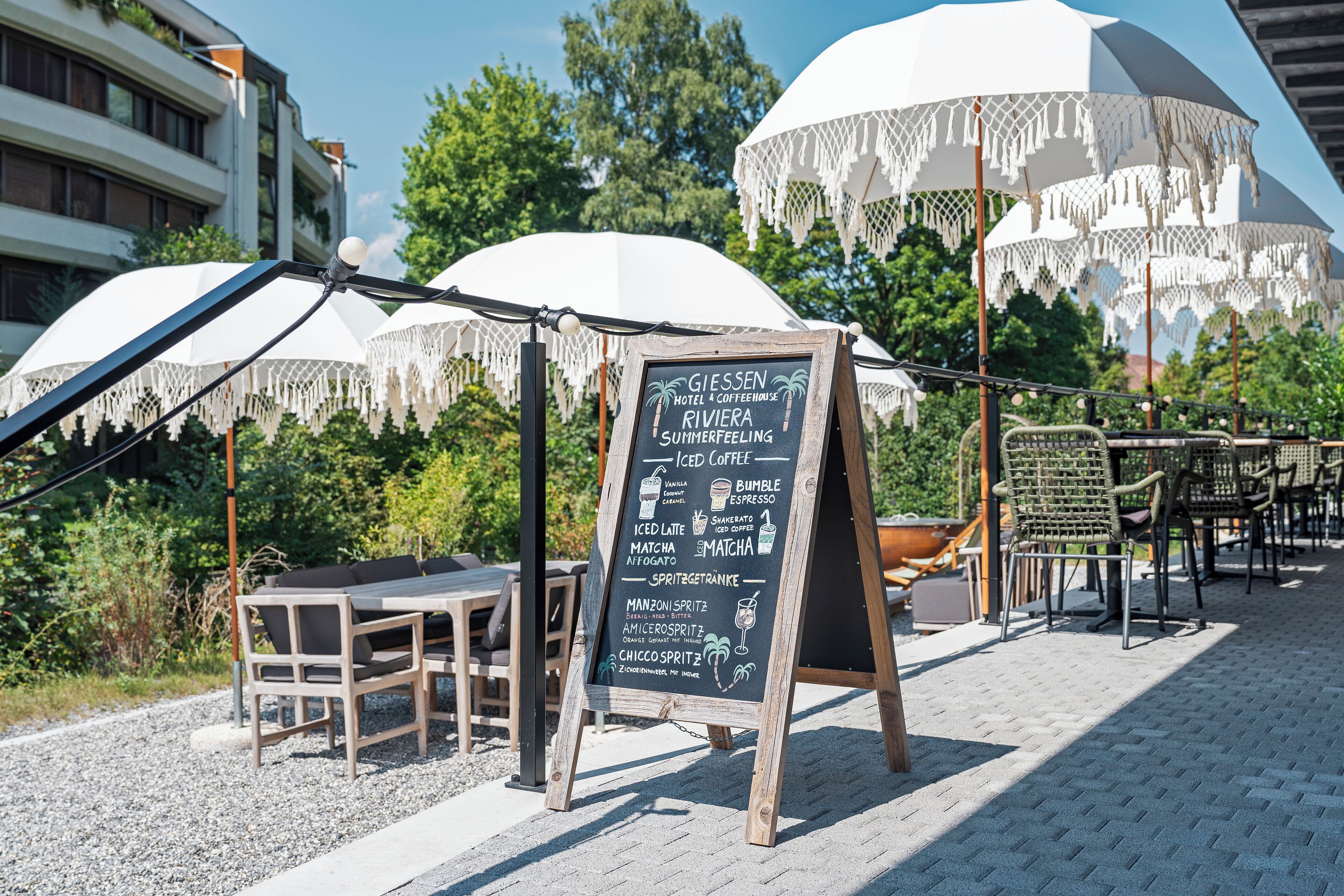 Un espace extérieur aménagé avec des tables et des chaises, un tableau noir servant de menu, des parasols et du mobilier de jardin.