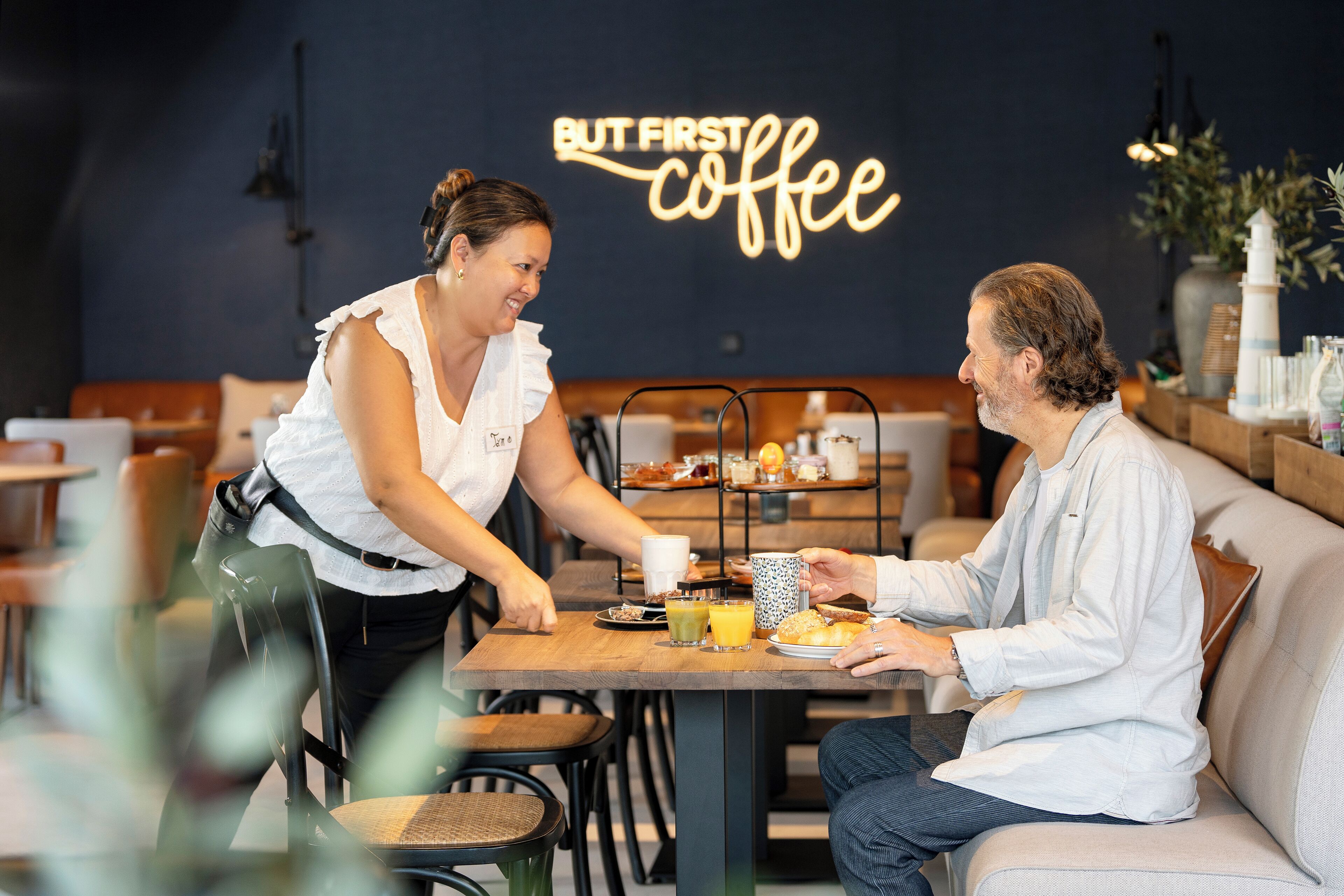 Una cafetería con un letrero de neón que dice "BUT FIRST COFFEE" y una mesa con una variedad de alimentos para el desayuno.