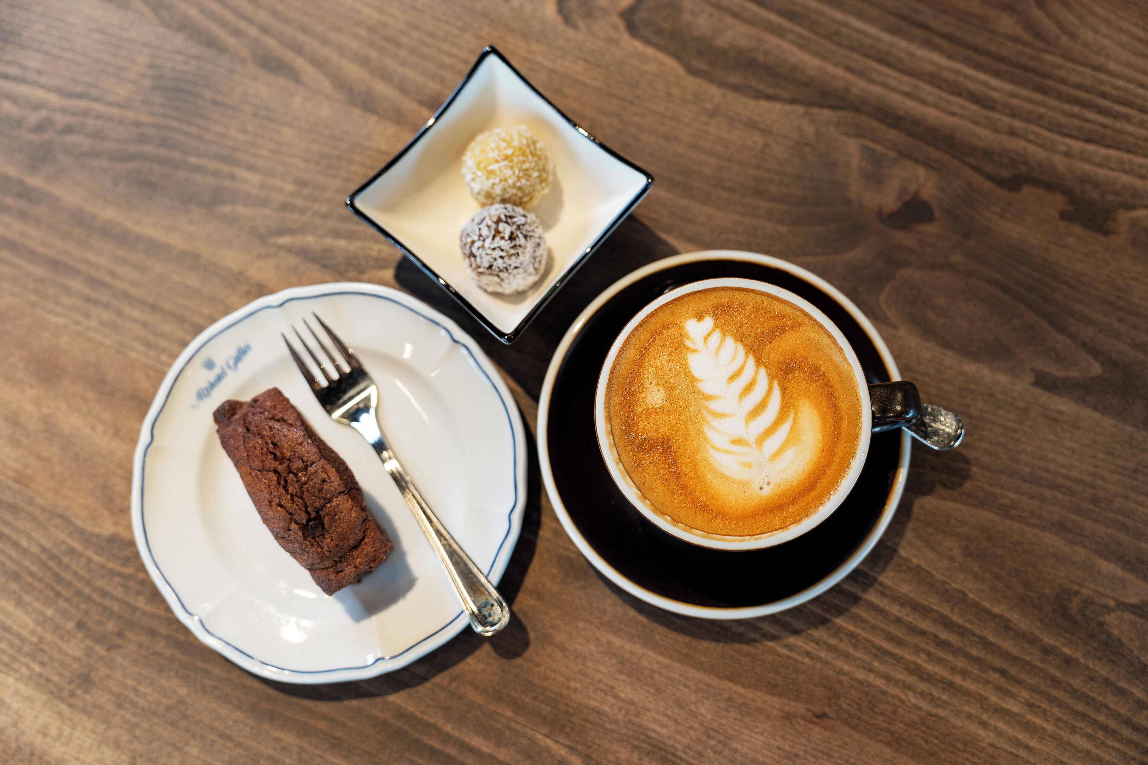 Un plato con un trozo de pastel y un tenedor, una caja pequeña con tres postres redondos, y una taza de café con arte latte sobre una mesa de madera.