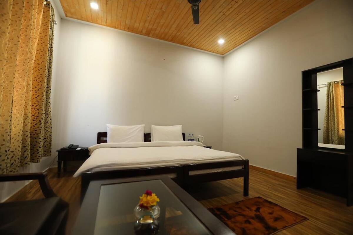 Hotellihuone, jossa on puinen katto, valkoisilla lakanoiden peitetty sänky, pieni pöytä maljakko täynnä kukkia ja peili seinällä.