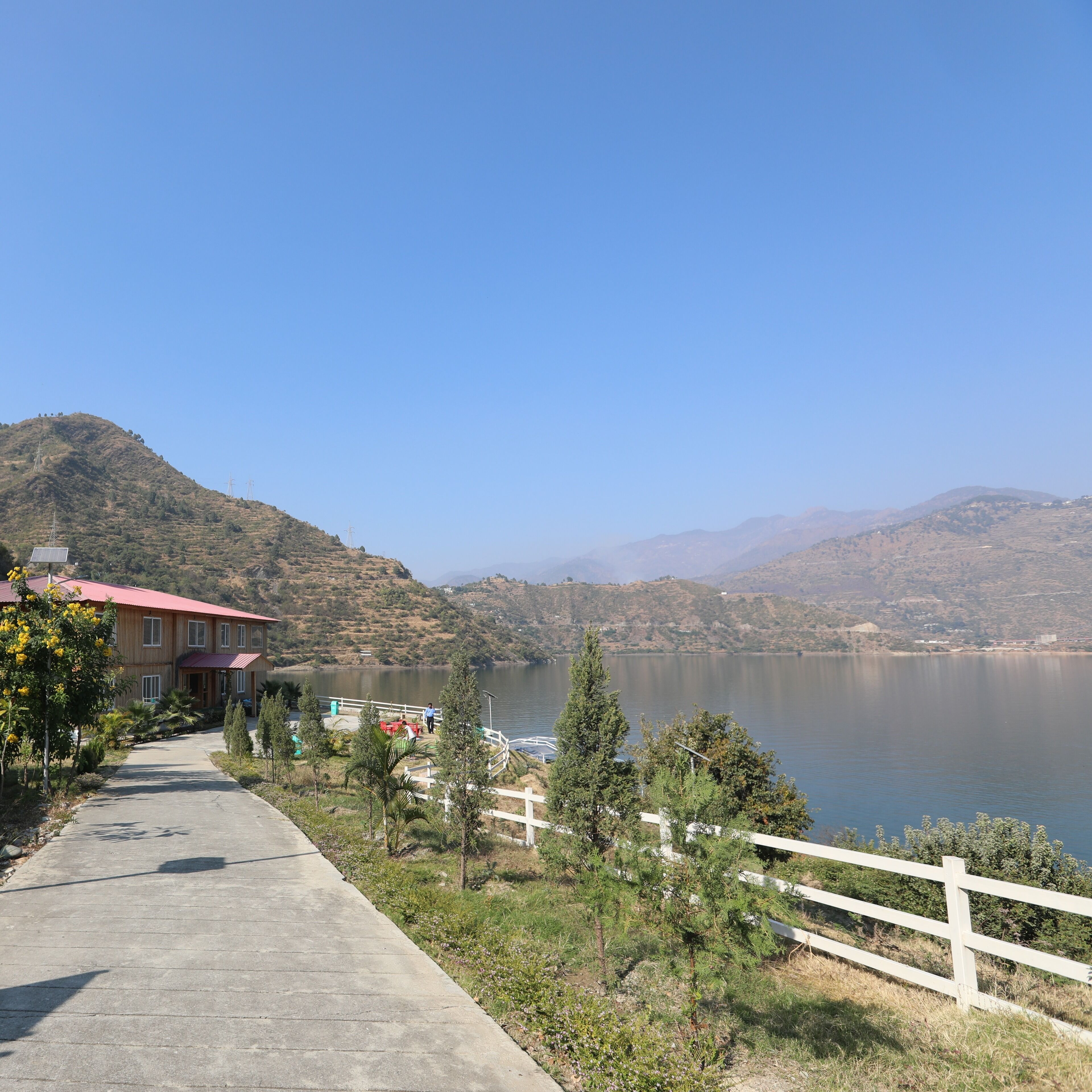 A lakeside view with a wooden building, a paved walkway, and a white fence.