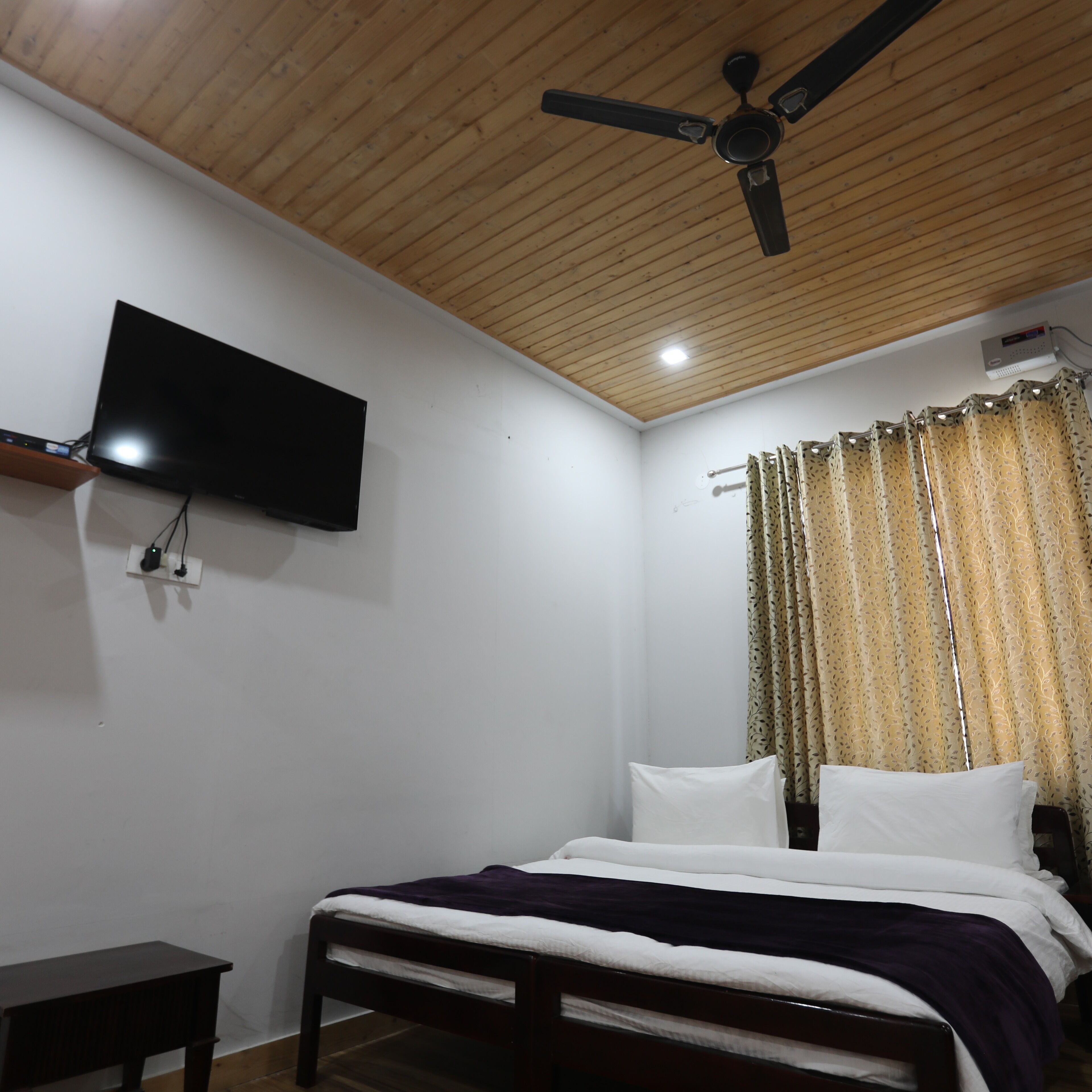 A bedroom with a bed, a ceiling fan, a TV mounted on the wall, and a curtain.
