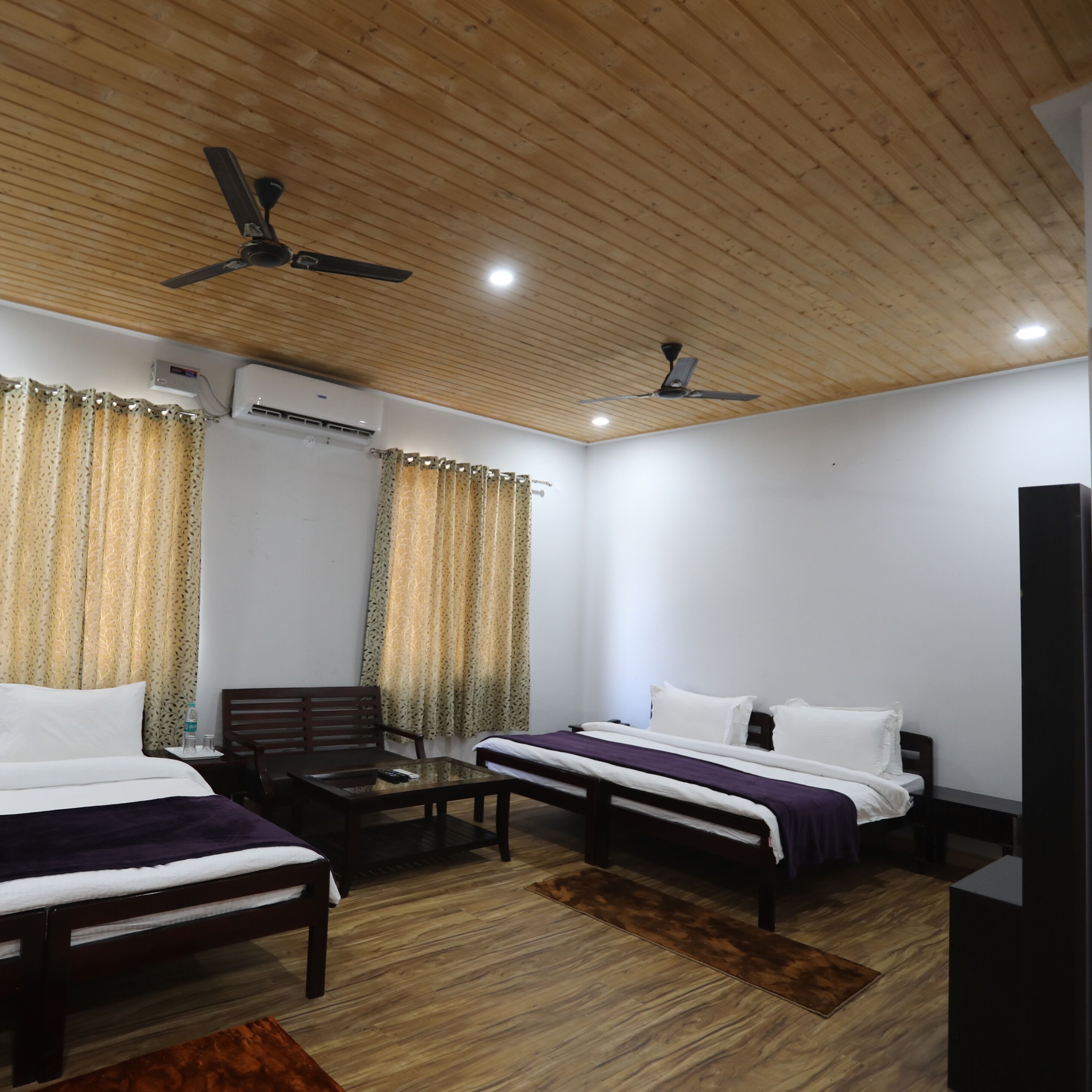 A hotel room with two beds, a wooden ceiling, ceiling fans, an air conditioning unit, and a television.