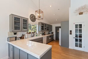 A modern kitchen with grey cabinets, a large island, a clock, and a hanging light fixture.
