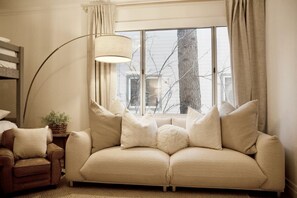 A cozy living room with a beige sofa, a small armchair, a floor lamp, and a window with a view of bare trees.