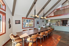 A spacious dining area with a long wooden table and leather chairs, a kitchen in the background, and a staircase leading to an upper level.