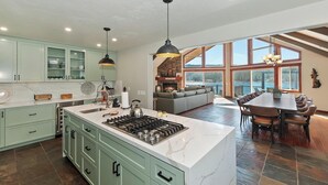 A spacious kitchen with a large island, a fireplace, and a dining area with a view of the lake. - It's a Wonderful Life by Destination Big Bear : Luxury Deep Water Lakefront! Pool Table! Panoramic Lake & Mountain Views! HOT TUB! (Big Bear Lake CA)