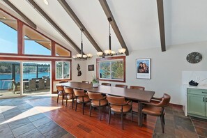 A spacious dining area with a large wooden table and chairs, a view of the outdoors through large windows, and a kitchen area with green cabinets.