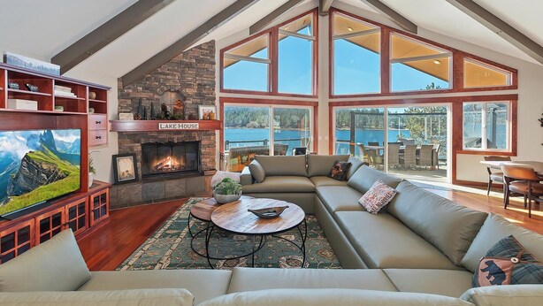 A spacious living room with a fireplace, large windows, and a view of a lake. - It's a Wonderful Life by Destination Big Bear : Luxury Deep Water Lakefront! Pool Table! Panoramic Lake & Mountain Views! HOT TUB! (Big Bear Lake CA)