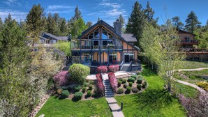 A large, multi-story house with a prominent balcony, surrounded by lush greenery and trees. - It's a Wonderful Life by Destination Big Bear : Luxury Deep Water Lakefront! Pool Table! Panoramic Lake & Mountain Views! HOT TUB! (Big Bear Lake CA)