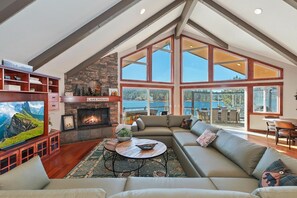 A spacious living room with a fireplace, a large window overlooking a body of water, and a television on a wooden stand.