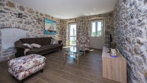 A living room with a stone wall, a brown sofa, a glass coffee table, and a floral ottoman. - Villa Dundo Pero (Mljet)