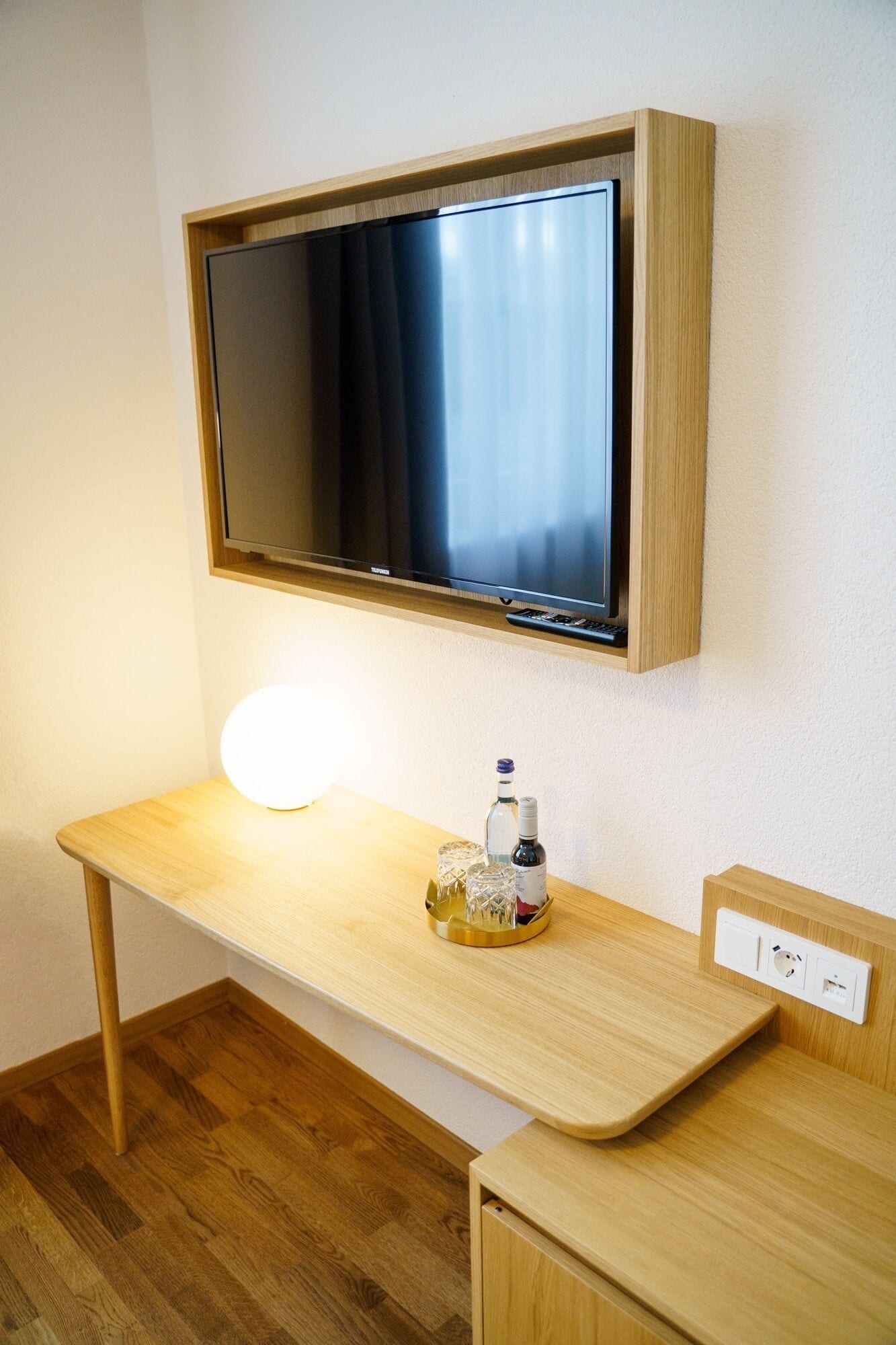 A hotel room with a wooden desk, a flat-screen TV mounted on the wall, a round table lamp, and a bottle of water.