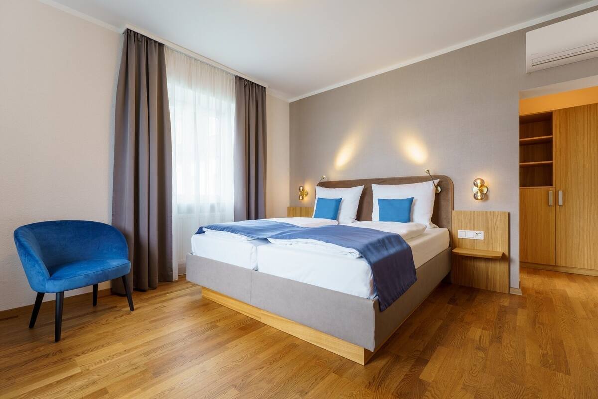 Una habitación de hotel con una cama, una silla azul, suelo de madera y un armario.
