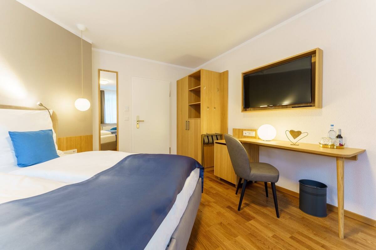 Una habitación de hotel con cama, escritorio, silla, televisor y cesto de basura.