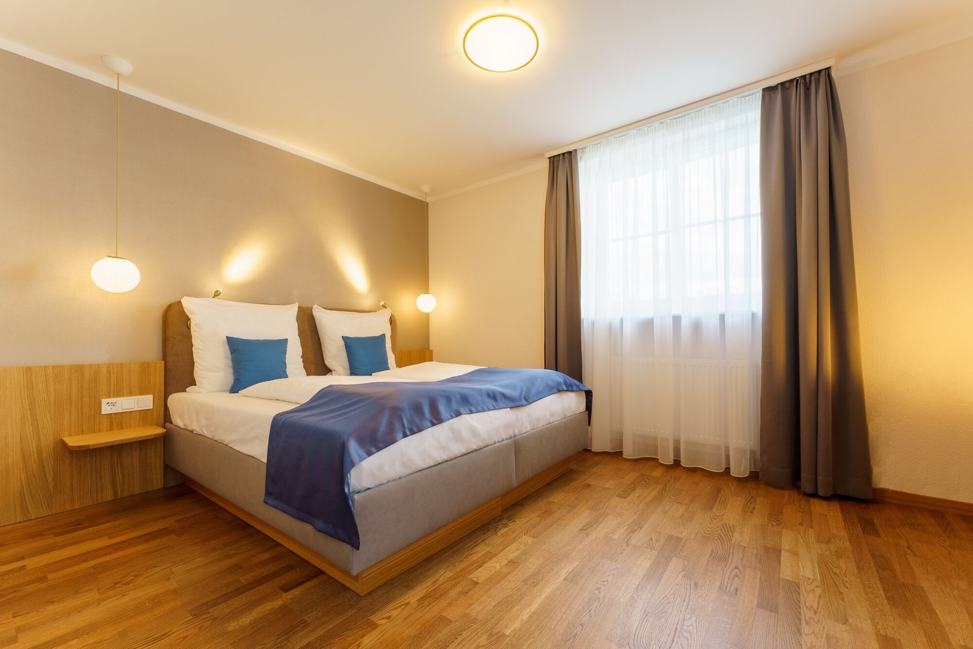 A modern hotel room with a wooden floor, a bed with white and blue bedding, two bedside tables, and a large window with curtains.