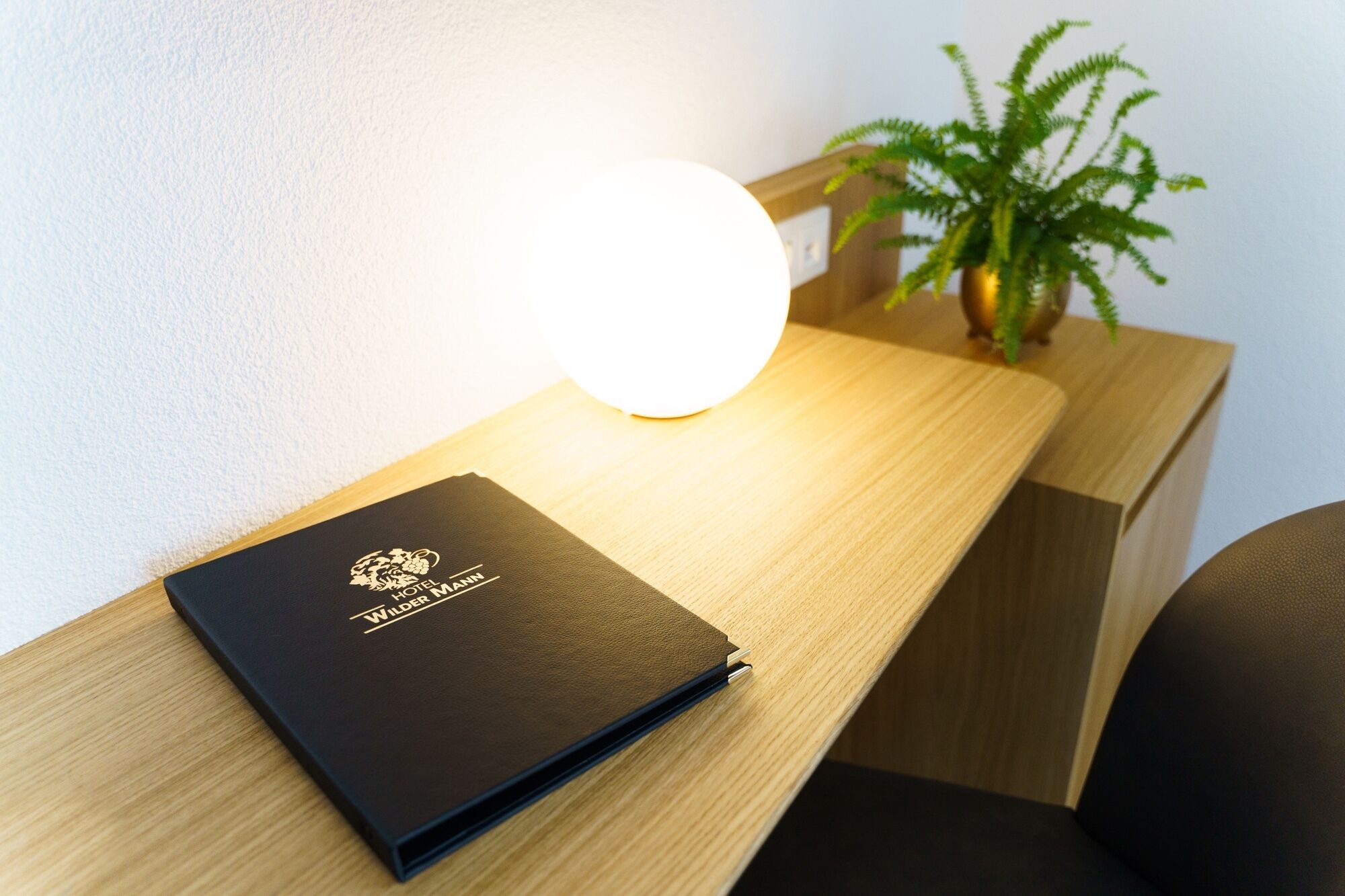 Ein Holztisch mit einem in schwarzes Leder gebundenen Buch mit dem Titel "Hotel Wilder Mann" und einer runden, beleuchteten Leuchte an der Wand.
