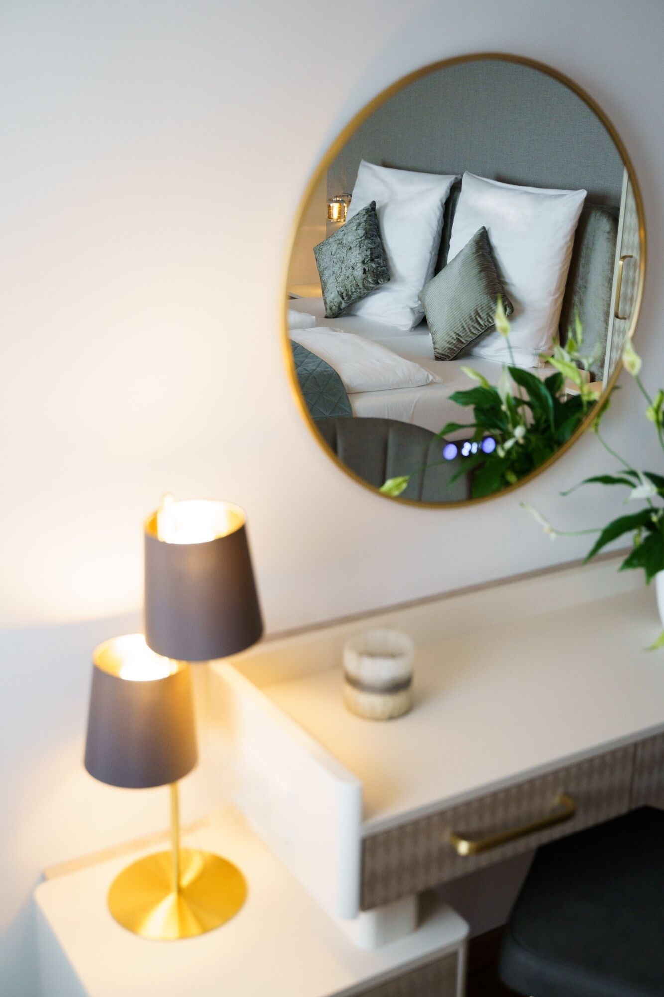 A bedside table with a lamp, a small vase, and a mirror reflecting a bed with pillows.
